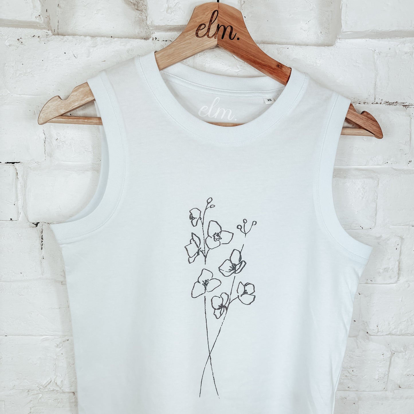 White tank top with floral design on a wooden hanger against a white brick wall.