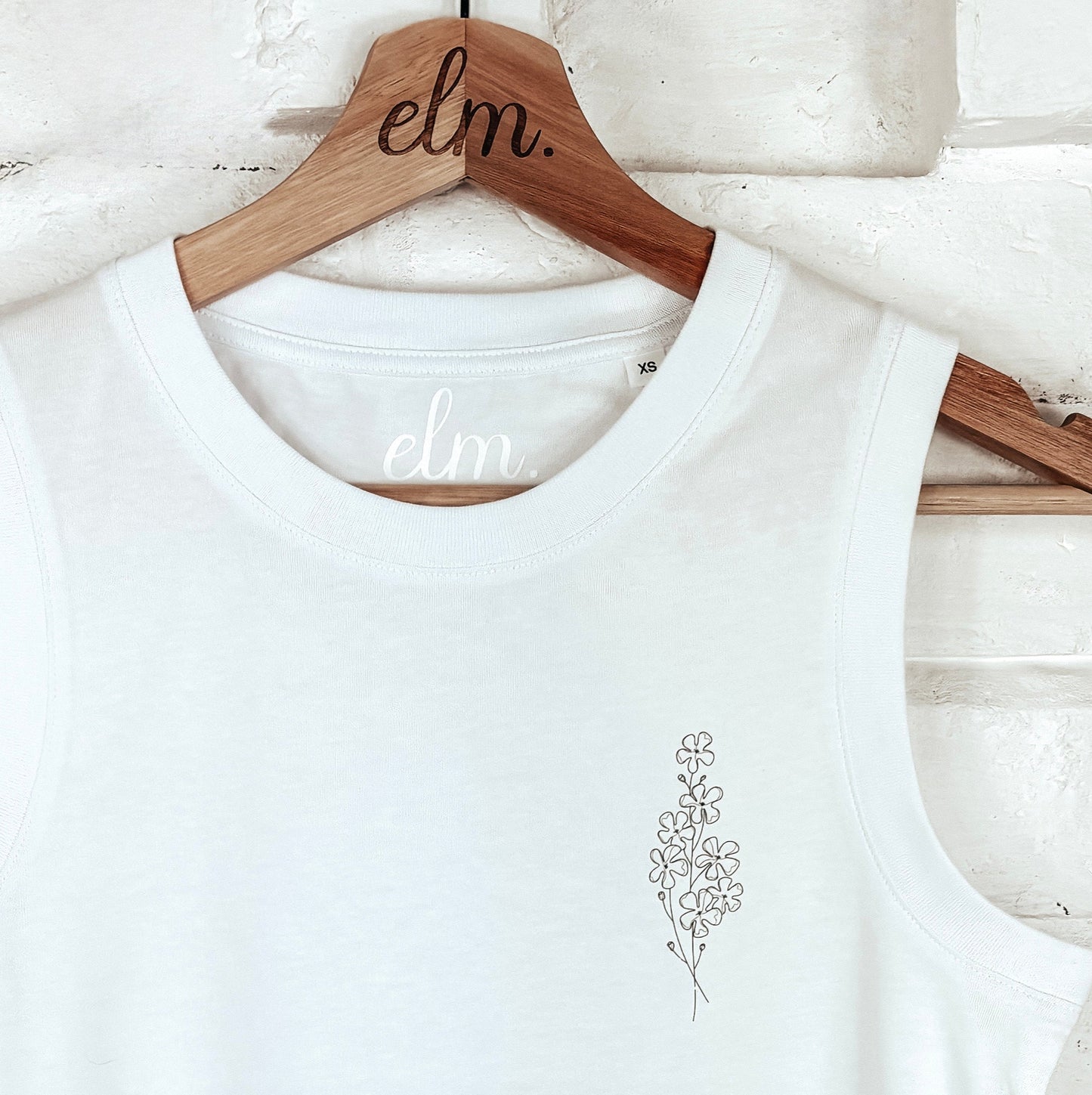 White tank top with a small design on a wooden hanger against a light brick wall.