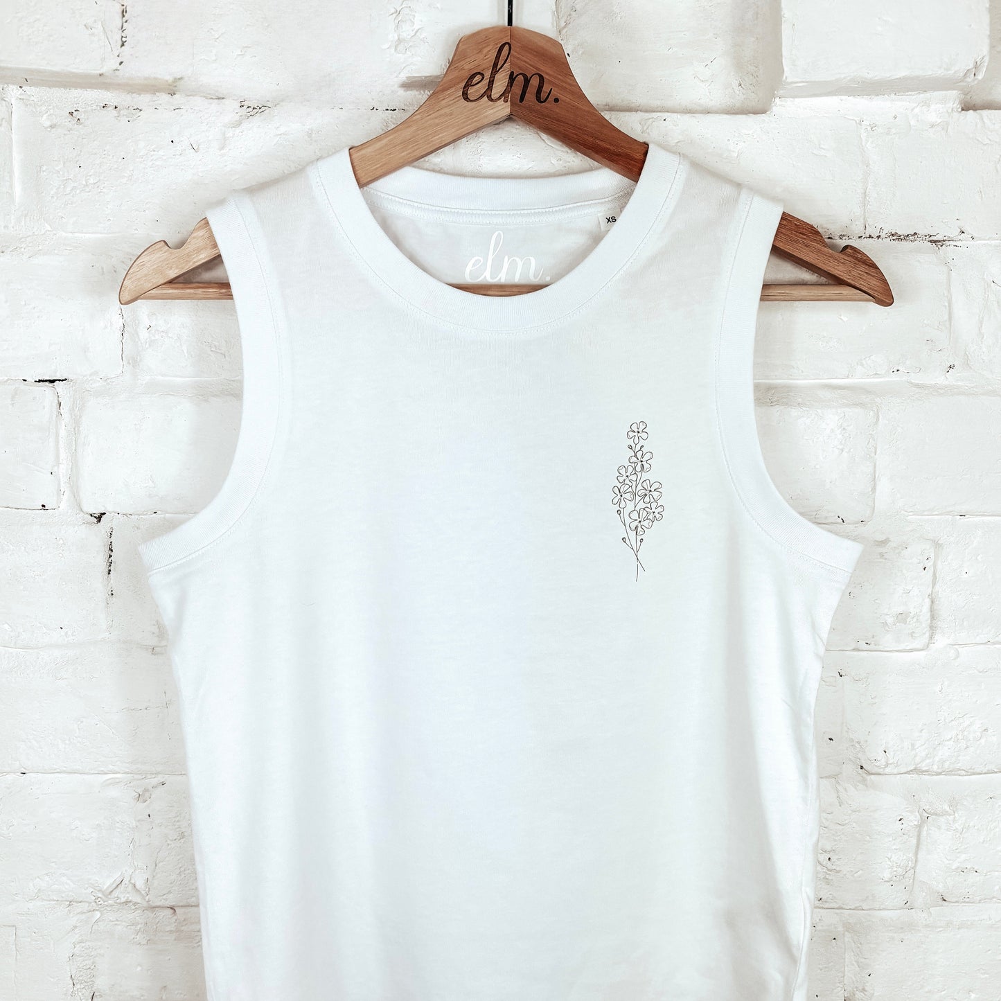 White tank top with a small design on a wooden hanger against a light brick wall.