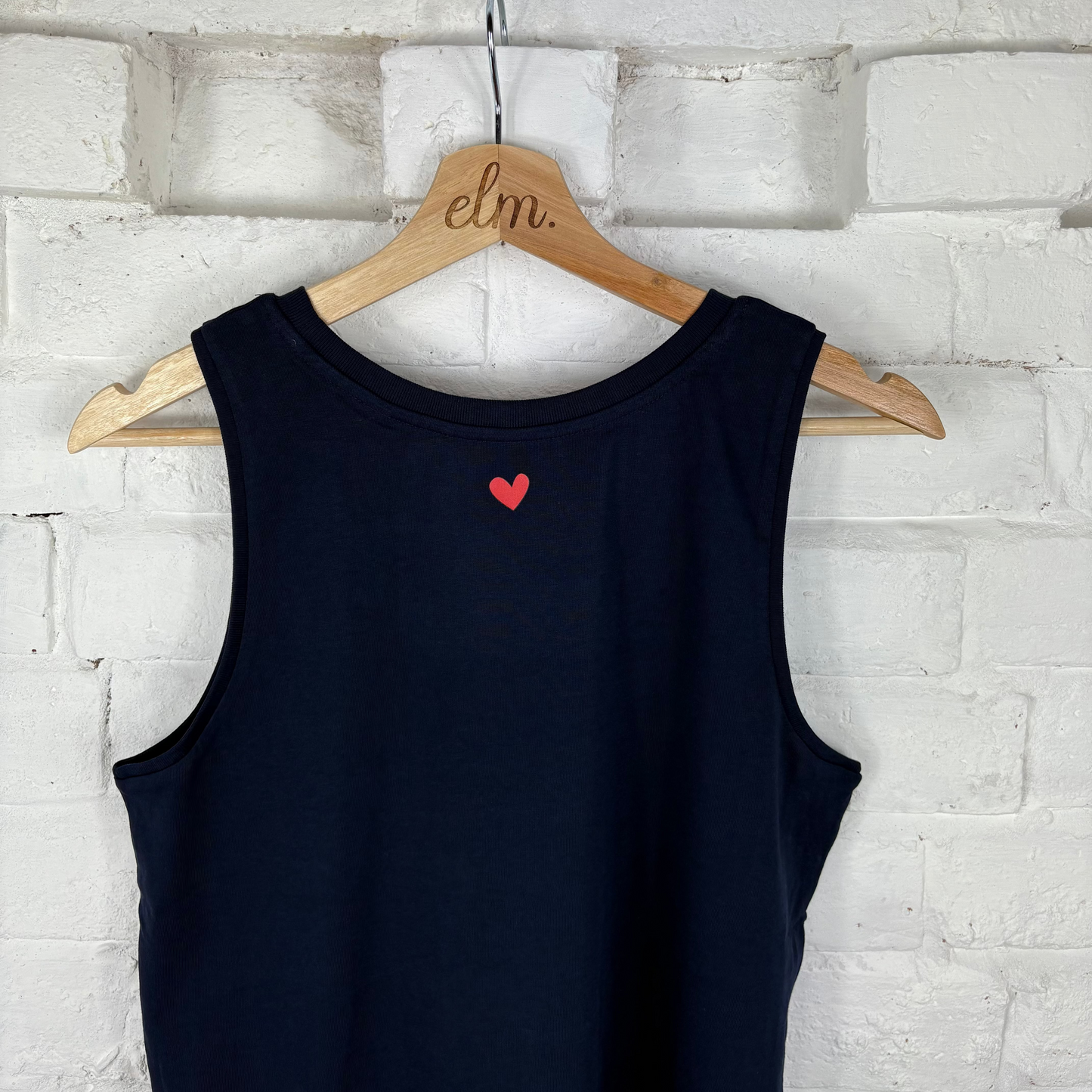 Navy  tank top with a red heart on a wooden hanger against a white brick wall.