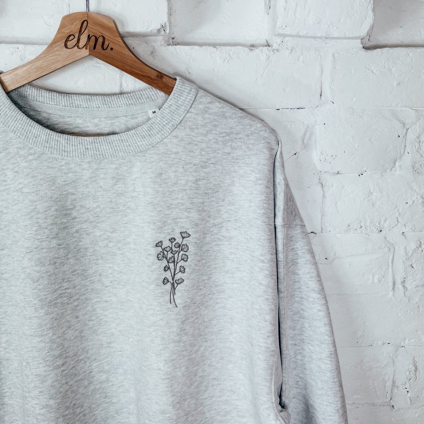 Gray sweatshirt with a small design on a wooden hanger against a white brick wall.