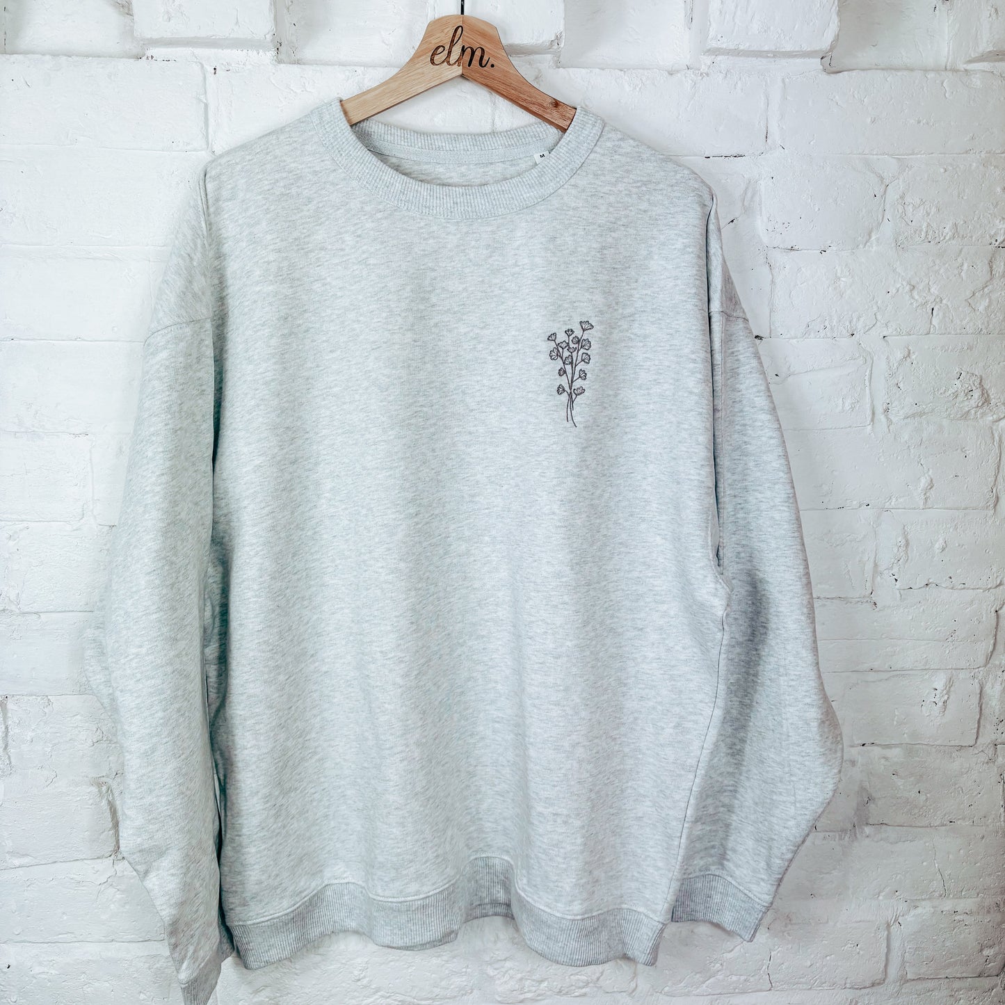 Gray sweatshirt with a small design on a wooden hanger against a white brick wall.